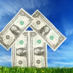 $1 bills arranged in the shape of a house on a grassy field against a blue sky.