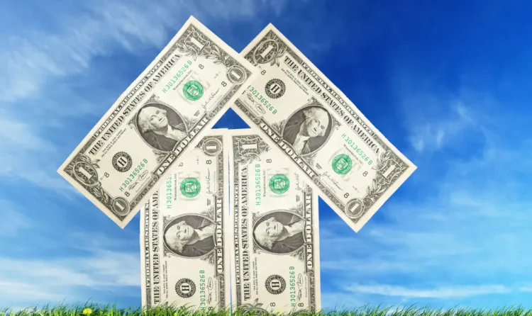 $1 bills arranged in the shape of a house on a grassy field against a blue sky.